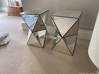 Two mirrored modern side tables placed on carpeted floor against a wall, showing the front and side triangular mirrored panels.