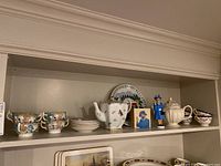 Shelf display showing floral teacups and saucers, white ceramic teapot, Queen Elizabeth II miniature, corgi portrait, decorative plate