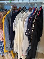 Photo showing a row of assorted women's clothes hanging on a rack including a bright yellow overcoat, white and striped shirts, dark cardigans, and tops.