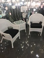 View of two white wicker armchairs with black cushions and matching square side table with glass top displayed on showroom floor