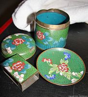 Photo of cloisonne cigarette tin, ashtray and matchbox holder with detailed floral pattern on green background, showing dent on tin rim.