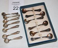 Photo showing two sets of silver salt spoons: one set of 6 in a blue fitted box and one set of 7 spoons laid out on table.