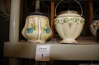 Two antique biscuit barrels side-by-side on a shelf, showing shape, color, and lids.