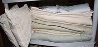 Stack of folded bed linens and two decorative pillows arranged on a shelf. Items include beige thermal-style blankets, sage green fleece blanket, quilted comforter, bed skirt, and two beige cutwork pillows.