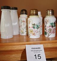 Five antique salt shakers displayed on a wooden shelf, four hand-painted opaque glass with floral designs and one pair of tall ribbed milk glass.