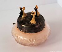 Vintage etched glass powder jar with metal lid featuring music box mechanism and three pairs of dancing figurines on top, photographed from a slight side angle