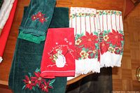 Photo showing five Christmas themed hand towels laid out on a wooden surface: two teal with poinsettia embroidery, one red with snowman embroidery, and two white with red poinsettia floral patterns and stripes with fringed edges.