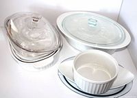 Photo showing the stack of white Corning Ware casserole dishes with clear glass lids and a round ribbed casserole dish in front with an oval glass casserole dish to the side.