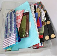 Photo displaying a plastic tub containing assorted colorful gift bags and tissue paper, including bags with stripes, florals, Christmas themes, and polka dots, as well as visible paper doilies.