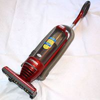 Photo of the Eureka branded red and gray bagless upright vacuum cleaner including dirt canister and flexible hose attachment.
