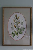 Photo shows an oval floral print with green leaves and small yellow flowers by Kovacs, professionally framed in a gold-colored frame with matting.