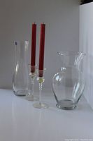 Two clear glass vases and two glass candlesticks with red candles placed on a white surface against a white backdrop.