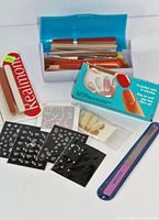 Photo showing a clear view of boxed emery boards, assorted packs of nail decorations with rhinestones and floral designs, a box of fake nails, a plastic pouch with fake nail tips, and a single purple emery board.