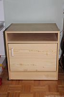 Front view of the wooden printer stand showing top surface, open middle shelf, and bottom cupboard with door and white handle. Light wood finish with visible grain patterns.