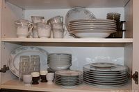 Full set of dishes and glassware arranged inside cabinet shelving, showing cups, plates, bowls, glass tumblers, and small egg cups.