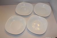 Four white shallow soup bowls with square rounded shape arranged on a flat surface.