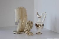 Three Greek decorative items arranged on a white surface against a light wall; includes the translucent Trojan horse book end, volcanic stone owl, and ceramic amphora