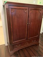 Closed front view of cherry-finished wooden armoire with two paneled doors, decorative shell carvings and full-width bottom drawer