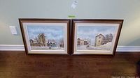 Two framed landscape paintings on canvas leaning against wall, showing snowy village scenes.