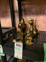 Brass decorative items including stylized cat and mouse figurines, an urn, mister, and vase, arranged on a black metal shelf with a pink wall background.
