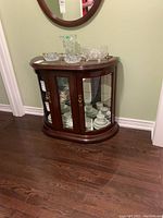 Front angled view of the wood cabinet with curved glass doors and shelves inside, placed against a light-colored wall, showing the polished wood finish and gold-tone handles.