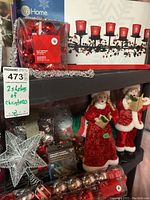 Two shelves of assorted Christmas decorations showing red candle holders, caroller figurines, ornaments, garlands, and star ornament.