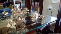 Four miniature clocks displayed on a glass shelf with decorative airplane, miniature lamp, and angel figurine. All items gold-toned and detailed.