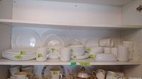 Shelves with various white Corelle dishes including plates, bowls, cups, butter dish, and creamer, organized in stacks with small green labels showing counts.