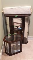 Small hexagonal wood and glass display case positioned in front of larger curved case, both on carpet, wrapped white packing material on top of larger case.
