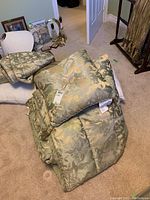 Photo of folded queen size comforter with matching throw pillows stacked on top, showing damask floral pattern in green and gold tones.