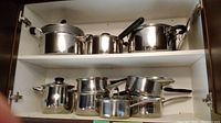 Nine stainless steel pots with lids arranged inside white cabinet shelving in two rows, showing various sizes and handle styles.