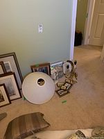 Overview photo of the grouped items showing framed art pieces leaning against the wall, lamp shades on the floor, and part of the figurine and lamps visible.