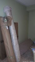 Photo of three large rolled rugs standing upright against a wall, two wrapped in brown paper and bubble wrap, one partially visible carpet leaning on side.