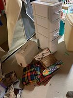 4-drawer plastic storage unit on wheels with two handles missing, various fabric pieces, fabric storage box, and cardboard sewing supplies box on floor in basement.