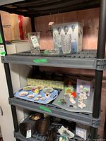 Gray plastic shelving unit with assorted decorative items including glass vases, ceramic figurines, small plates on tray, and a boxed light fixture. Located in basement with unfinished walls visible.