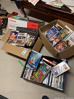 Two open cardboard boxes on basement floor containing Toshiba combo player box and multiple VHS/DVD cases