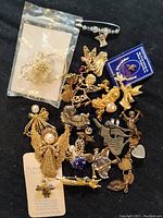 Top-down photo showing assorted angel and cherub costume jewelry brooches on black background, including gold-tone filigree angels, silver-tone pieces, and some in original packaging.