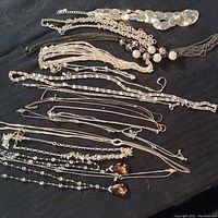 Overview of assorted silver tone Avon necklaces arranged on black fabric, showing variety of chain types, bead embellishments, and pendants.