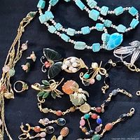 Display of assorted Egyptian revival style jewelry on black fabric, showing necklaces, bracelets, earrings, and brooches with carved semi-precious stones including scarabs and butterfly motifs.