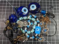 Collection of various evil eye jewelry pieces on black grid background showing pendants, bracelets with beads, leather strap with metal angel charm and gold-tone studs, and keychains.