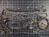 Top view of assorted gray cultured pearl necklaces and bracelets laid out on a grid background, showing varying sizes of pearls and different chain styles.
