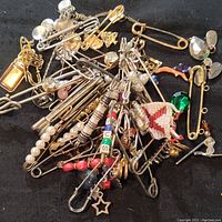 Top view of assorted oversized safety pin kilt pins piled together showing variety of decorative elements including beads, charms, and woven pieces.