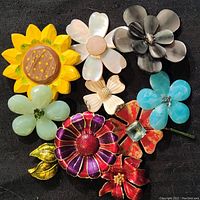 Full view of all eight assorted oversized flower brooches arranged on a black background highlighting the variety of colors and materials including carved mother of pearl petals, enamel, and metal.