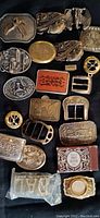 Top-down photo showing 21 assorted men’s belt buckles on a dark surface, various shapes and designs including animals and brand logos.