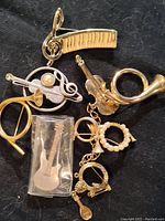 Various assorted musical instrument shaped brooches placed on a black background showing details of piano keyboard, violin, French horn, and pearl accents.