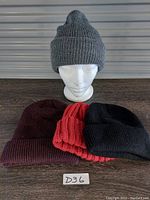 Four ribbed knit winter toques in red, burgundy, black, and grey. Grey toque displayed on mannequin head, others flat.