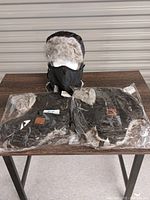 Photo showing two black fuzzy toques with gray faux fur lining on ear flaps and face masks displayed on table. Both are still in packaging.