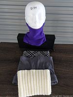 Photo shows 4 neck warmers: purple knit worn on mannequin head, gray fleece folded, plaid pattern neck warmer partially visible under fleece, and cream ribbed knit neck warmer folded on table.