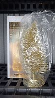Gold 10 inch glitter Christmas tree wrapped in clear plastic sitting in front of its box, showing the tree's shape and gold glitter finish.