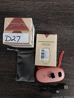 Photo showing one pink Unigear rechargeable hand warmer, two boxes with product labeling and barcodes, black soft pouch, and a USB charging cable, all on dark wood surface.
